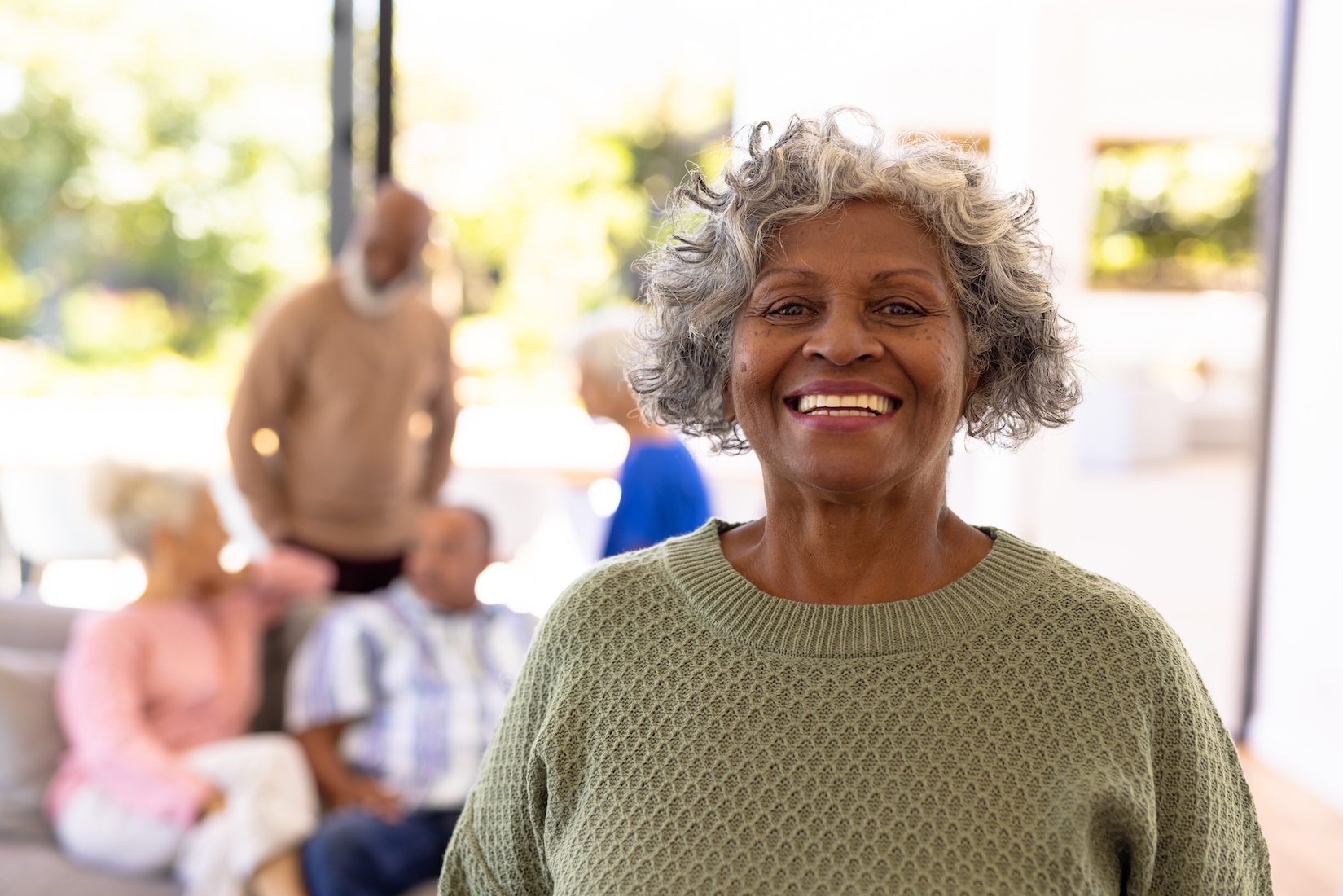 Senior woman in sage green sweater stands smiling at the camera with multiple friends talking together in the background.